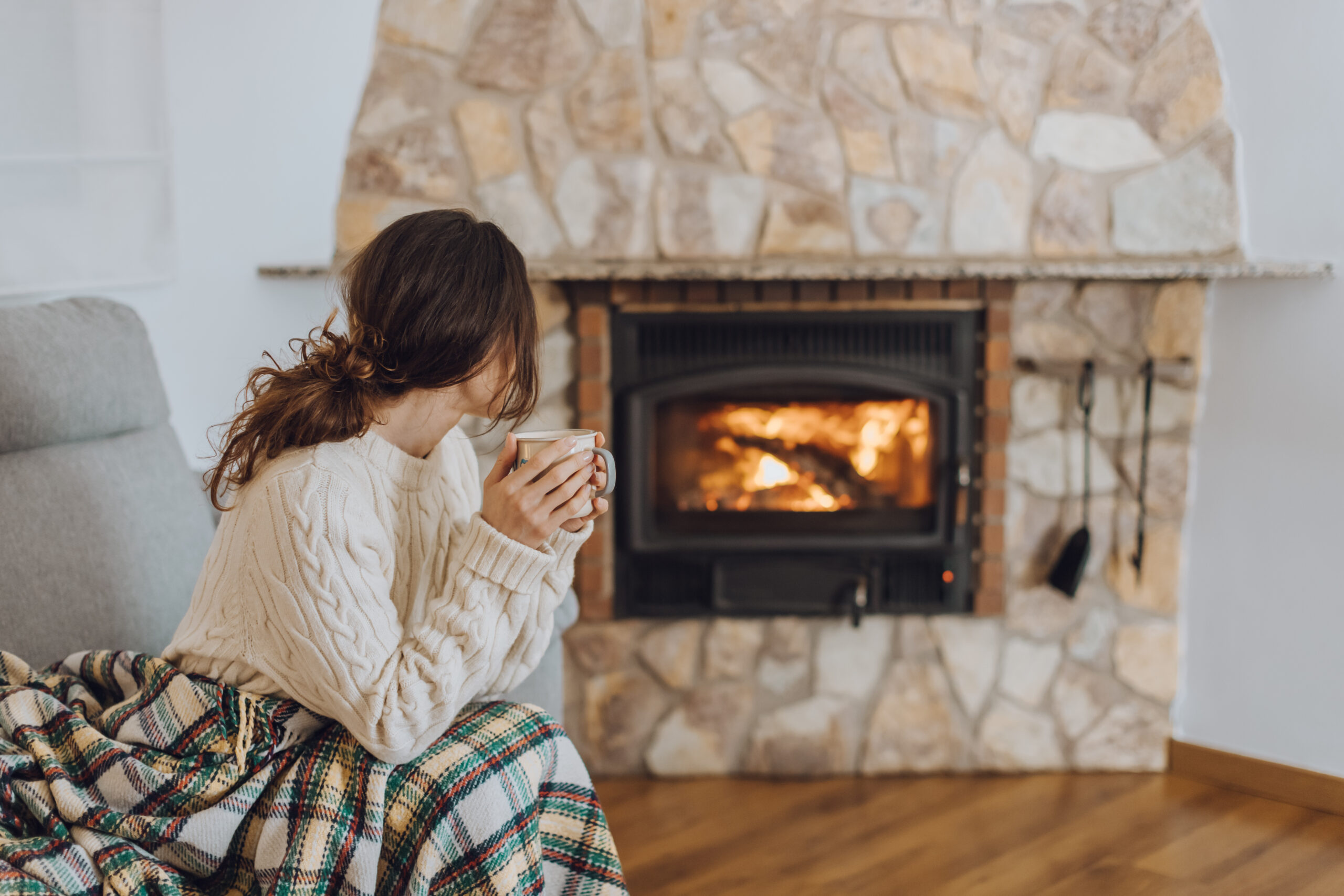 Adobe stock image of a woman sitting by a stone fireplace with a warm drink.